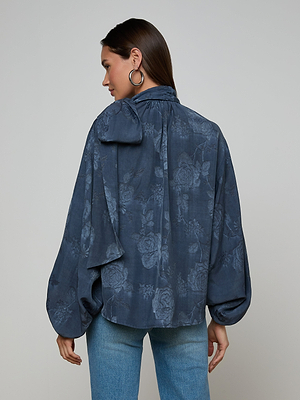 A woman faces away, wearing the Rhett Oversized Blouse in dark blue floral jacquard with voluminous sleeves and a large neck bow, paired with light blue jeans. Her brown hair is side-parted and she accessorizes with large hoop earrings.