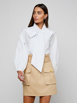 A woman wears the Deon Tie-Neck Blouse with blouson sleeves in organic cotton poplin, paired with a high-waisted beige skirt with oversized front pockets and gold buttons, posing against a plain light gray background.