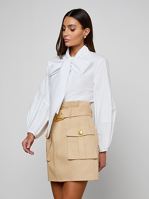 A woman wears the Deon Tie-Neck Blouse in organic cotton poplin with blouson sleeves, paired with a high-waisted beige skirt with large pockets and gold buttons, set against a plain light gray background.