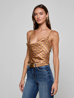 A woman with long brown hair wears the L'AGENCE Calista Silk Camisole in a gold and red horse bit print, paired with blue jeans, a tan belt, and gold hoop earrings, standing against a plain light gray background.