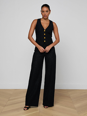 A woman poses against a plain light wall, wearing the Kino Lace Vest, a sleeveless black jumpsuit with gold buttons, modern tailoring, wide-leg pants, and black heeled sandals. Her hands are clasped in front.