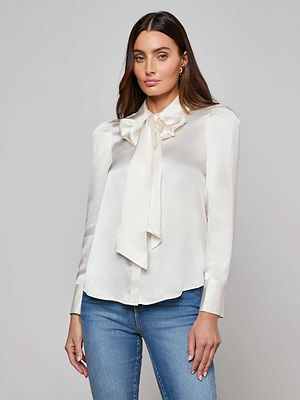 A woman with long brown hair and a neutral expression wears the Honey Silk Blouse, featuring a large bow at the neck, along with blue jeans against a plain background.