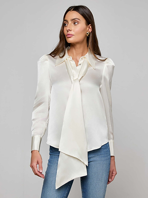 A woman wears the Honey Silk Blouse in cream silk charmeuse with a tie neck and long sleeves, paired with blue jeans, standing against a plain light gray background.