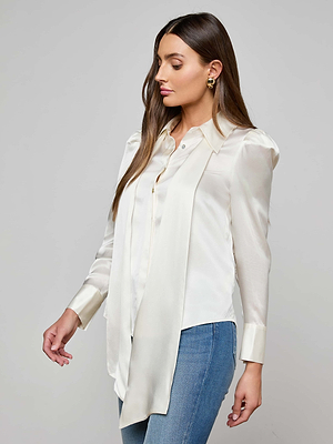 Woman with long brown hair wears the Honey Silk Blouse, a cream silk charmeuse top with tie neck and scarf detail, paired with gold hoop earrings and blue jeans, standing against a plain light gray background.