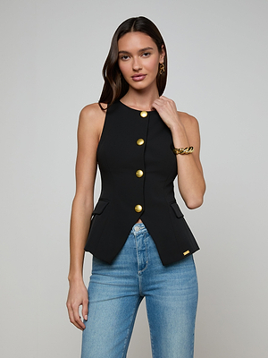 A woman with long brown hair models the Tessa Vest, a sleeveless suit vest with gold buttons, paired with light blue jeans. She poses against a plain gray background and accessorizes with gold hoop earrings and a gold bracelet.
