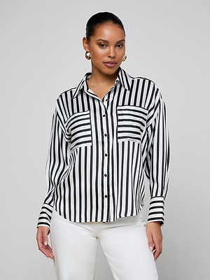 A woman wearing the Eliora Shirt—a black and white striped button-down with chest pockets—and white pants poses confidently against a plain light gray background.