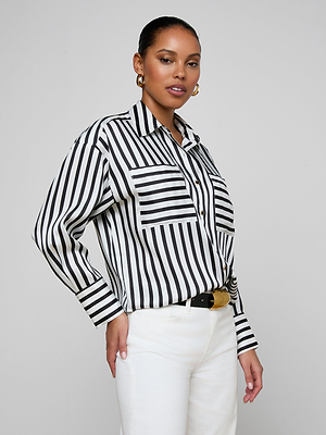 A woman wears the Eliora Shirt, a black and white vertical striped button-down with a relaxed fit, paired with white pants and gold hoop earrings, standing against a light gray background and smiling slightly at the camera.