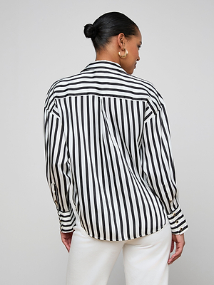 A woman with her hair in a low bun stands with her back to the camera, wearing the relaxed fit Eliora Shirt featuring black-and-white stripes, paired with white pants against a plain background.
