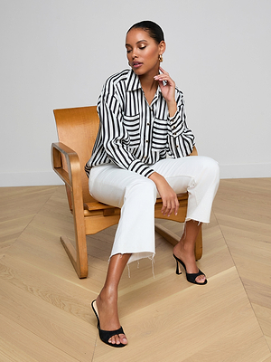 A woman sits on a wooden chair in a modern minimalistic room, wearing the Eliora Shirt— a relaxed fit striped button-down— paired with white frayed-hem pants and black heeled sandals, posing with one hand touching her face.