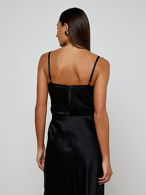 A woman with straight dark hair wears the Orlina Silk Camisole—a sleeveless black silk charmeuse top with thin straps—seen from behind against a plain light background. She accessorizes with round gold earrings.