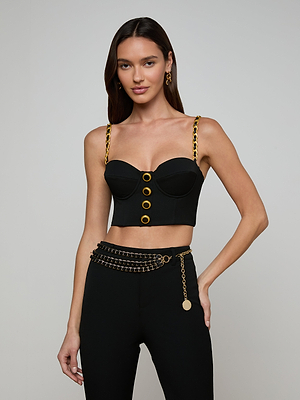 A woman wears the Sophia Chain Bralette in black with gold chain hardware and buttons, styled with black pants featuring a gold chain belt, gold earrings, long brown hair, and a neutral expression against a plain light gray background.