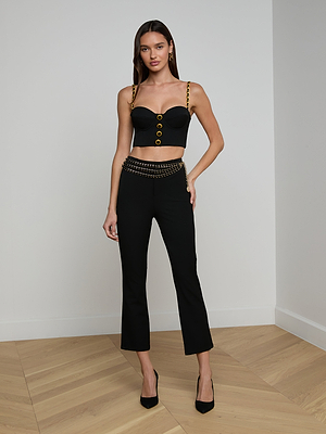 A woman stands in a corner of a room with light wood floors, wearing the Sophia Chain Bralette with gold chain details, black stretch suiting pants with gold chain accents at the waist, and black heels.