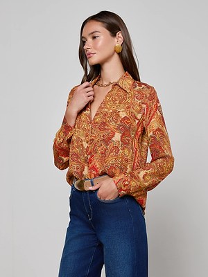 A woman stands against a plain background wearing the Nina Silk Blouse by L'AGENCE, a relaxed-fit, golden paisley silk button-down with warm red and gold tones, paired with blue jeans, gold earrings, and a gold necklace.