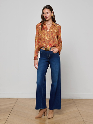 A woman stands indoors wearing the L'AGENCE Nina Silk Blouse in a golden paisley print with a relaxed fit, styled with blue wide-leg jeans, beige heeled shoes, a brown belt, and accessorized with earrings and a necklace.