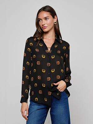 A woman wears the L'AGENCE Nina Silk Blouse, a relaxed fit black button down with a yellow and orange geometric buckle print, paired with blue jeans against a plain light gray background.
