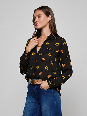 A woman with long brown hair wears the L'AGENCE Nina Silk Blouse—a relaxed-fit black button-down with a yellow and brown pattern—paired with blue jeans and gold earrings. She stands touching her collar, looking to the side against a plain background.