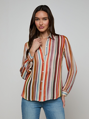 A woman with long brown hair wears the Nina Silk Blouse, a relaxed-fit, multicolored stripe button-up shirt, paired with blue jeans. She stands against a plain light background, gently touching her collar and smiling slightly.