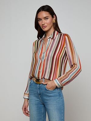 A woman wears the Nina Silk Blouse—a relaxed fit, multicolored stripe long-sleeve shirt—with light blue jeans and large gold hoop earrings, standing against a plain light background with one hand in her pocket.
