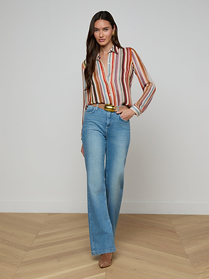 A woman stands indoors on a wooden floor, confidently posing in front of a white wall wearing the Nina Silk Blouse—a relaxed fit, multicolored striped top—paired with light blue high-waisted jeans, tan heels, and a yellow belt.