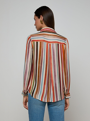 A woman with straight brown hair and gold statement earrings faces away, wearing the Nina Silk Blouse—a relaxed-fit, multicolored stripe shirt—paired with blue jeans against a plain light background.
