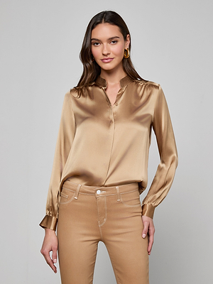 A woman with long brown hair wears the Bianca Silk Blouse in shiny gold and tan pants, standing against a plain gray background and smiling softly at the camera.