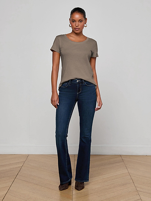 A woman stands indoors against a white wall, wearing the L'AGENCE Cory Cotton Scoopneck Tee in taupe, dark blue bootcut jeans, brown heeled shoes, and gold hoop earrings. Her hair is pulled back and she has a neutral expression.