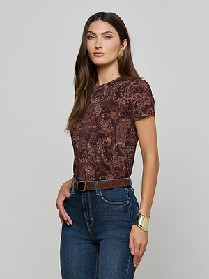 A woman with long brown hair wears the L'AGENCE Ressi Fitted Tee with a paisley print, blue jeans and brown belt, statement earrings, and a gold bracelet, standing against a plain light gray background.