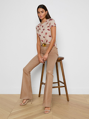 A woman with long dark hair, wearing the bestselling Ressi Fitted Tee in stretch jersey with a lips print, tan slim cut flared pants, and heeled sandals, sits on a wooden stool against a white wall and light wood floor.