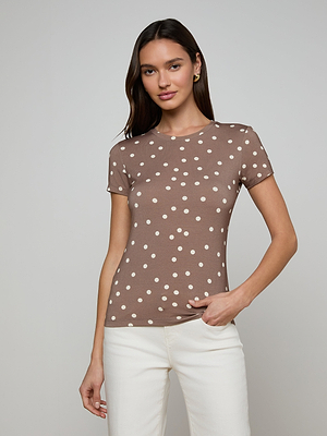 A woman with long dark hair smiles softly while wearing the Ressi Fitted Tee—a brown, slim cut, short-sleeve shirt with white polka dots—and white pants, standing against a plain background with one hand in her pocket.