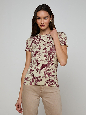 A woman with long brown hair stands against a plain backdrop wearing the Ressi Fitted Tee in beige with a maroon butterfly toile print and matching beige pants, looking relaxed as she touches her neck.