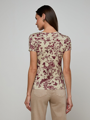 A woman with long brown hair faces away, wearing the Ressi Fitted Tee—a slim-cut beige short-sleeve top with a maroon floral pattern—and light beige jeans against a plain light background.