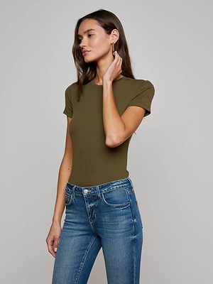 A woman wearing the L'AGENCE Ressi Micro-Rib Tee in olive green and blue jeans stands against a plain background, touching her hair with one hand and gazing to the side.
