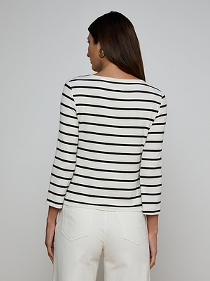 A woman stands facing away, wearing the Lucille Striped Boatneck Top with classic stripes and a boat neckline, paired with white high-waisted pants. She completes her look with long straight brown hair and gold hoop earrings.