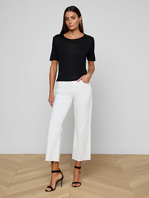 A woman with long brown hair stands indoors against a plain light wall, wearing the black Remy Tee in lightweight jersey, white wide-leg cropped pants, and black heeled sandals. Her hand rests in her pocket.