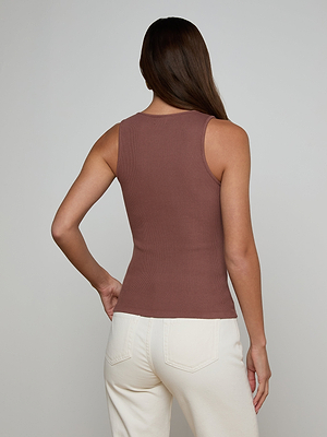 A woman with long brown hair, seen from behind, wears the Adira Scoopneck Tank and off-white pants. She stands against a plain light gray background with one hand on her hip.