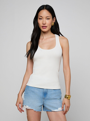A woman with long black hair wears the Adira Scoopneck Tank in white, paired with light blue denim shorts, gold hoop earrings, and bangles, standing against a plain gray background.