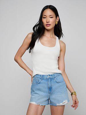 A woman with long black hair wears the Adira Scoopneck Tank and light blue ripped denim shorts. She stands against a plain light gray background, accessorized with gold earrings, a bracelet, and one hand on her hip.