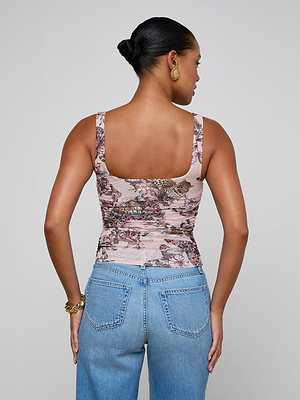 A woman with her hair in a bun stands with her back to the camera, wearing a sleeveless Rasha Shirred Mesh Tank in butterfly-jungle print and light blue jeans, accessorized with gold hoops and a bracelet against a plain background.