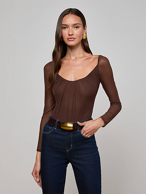 A woman with long brown hair wears the Gwendolyn Mesh Bodysuit, featuring sheer long sleeves, paired with dark blue jeans, a brown belt with a large gold buckle, and gold earrings, standing against a light grey background.