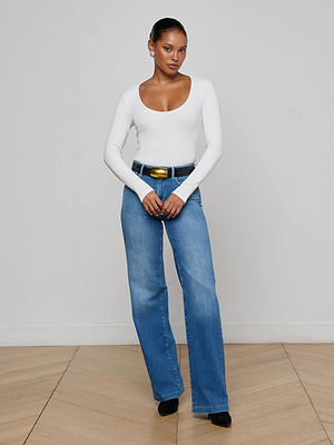 A woman stands indoors against a light wall, wearing the Scarlet Scoopneck Tee by L'AGENCE, paired with blue jeans, a black belt, and black shoes. Her hair is pulled back and she stands on a light wood floor.