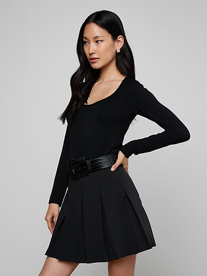 A woman with long dark hair wears the Cassandra Ruched Bodysuit, a fitted black long sleeve piece with a scoop neckline, paired with a pleated skirt and wide belt. She stands sideways, hand on her hip, against a plain light gray background.