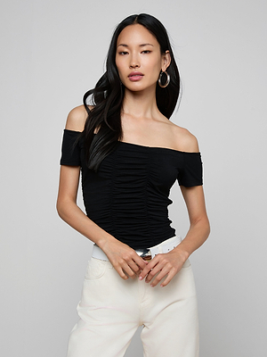 A woman with long black hair poses confidently in the Baliegh Ruched Bodysuit—an off-the-shoulder black rayon stretch jersey top with allover ruching—paired with white jeans, against a plain light gray background.