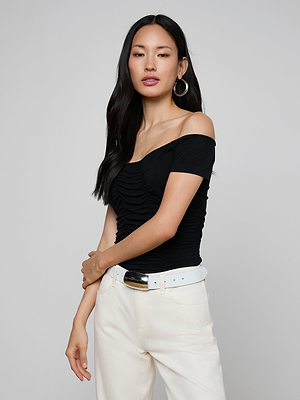 Woman with long black hair, large hoop earrings, and a white belt wears the Baliegh Ruched Bodysuit—an off-the-shoulder black rayon stretch jersey top with allover ruching—paired with white pants, against a plain light gray background.