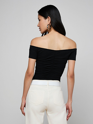 A woman with long dark hair is seen from the back in the Baliegh Ruched Bodysuit, a black off-the-shoulder ruched top, paired with high-waisted white pants and large hoop earrings, standing against a gray background.