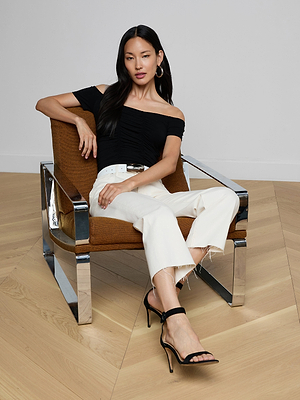 A woman with long dark hair poses confidently in a brown chair, wearing the Baliegh Ruched Bodysuit—an off-the-shoulder black rayon stretch jersey top—paired with white frayed-hem pants, a white belt, and black high-heeled sandals.