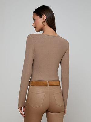 A woman with long brown hair wears the Vanya Scoopneck Top as a fitted base layer, paired with high-waisted beige pants and a brown belt, facing away from the camera against a plain light background.