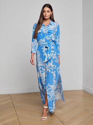 A woman with long brown hair stands indoors wearing the Cameron Silk Shirt Dress, a long-sleeved blue maxi dress featuring a white paisley print, tied waist belt, and paired with white high-heeled sandals.
