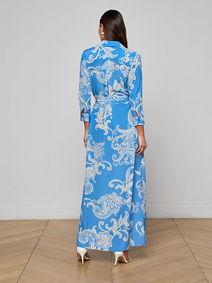 A woman stands indoors on a wooden floor, facing away, wearing the Cameron Silk Shirt Dress—a long blue paisley-print silk dress with three-quarter sleeves—and white high heels, against a plain light-colored wall.