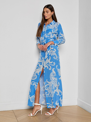 A woman indoors wears the Cameron Silk Shirt Dress, a blue long-sleeve maxi with a white paisley print and front slit. She has long straight hair and pairs it with white high-heeled sandals.