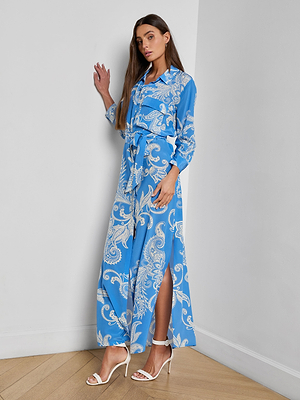 A woman stands indoors against a white wall, wearing the Cameron Silk Shirt Dress—a long-sleeved blue maxi dress with a white paisley print, waist tie, and high slit. She styles it with white heeled sandals and her long brown hair loose.